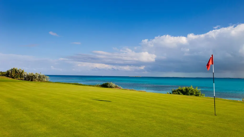 A golf course in Guanacaste with a red flag on a green, overlooking the calm blue sea under a partly cloudy sky.